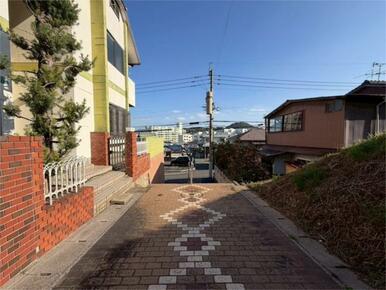 青空が広がる日差し明るい現地。前面道路は舗装されており、住宅街の中を穏やかに下っていく坂道です。