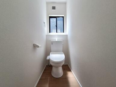 明るい窓付きのトイレ空間です。白を基調とした内装で清潔感があります。床は木目調で落ち着いた雰囲気です。換気がしやすく快適にご利用いただけます。無駄のないシンプルな設計です。