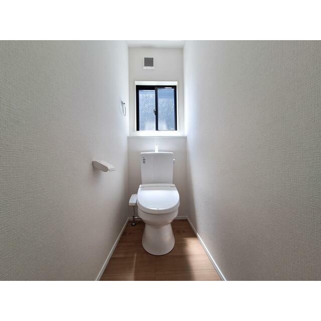 明るい窓付きのトイレ空間です。白を基調とした内装で清潔感があります。床は木目調で落ち着いた雰囲気です。換気がしやすく快適にご利用いただけます。無駄のないシンプルな設計です。